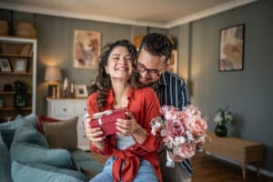 Young man surprising his girlfriend with a gift and flowers at home
