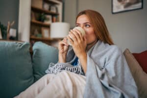 Cozy portrait of a sick mid adult woman wrapped in a blanket, enjoying a warm drink on the sofa in her living room, finding comfort and relief