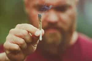 Young bearded man holding a smoking marijuana joint - legalization medical cannabis concept - smoke is coming out of the weed or hashish cigarette