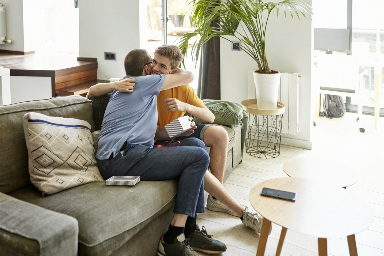 Full length view of mature man and teenage boy sitting together on sofa in family home and opening gift. Part of lifestyle series depicting deaf family and friends.