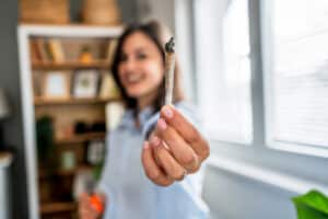 Smiling young woman holding a marijuana cigarette while relaxing in her cozy living room, enjoying a moment of leisure at home
